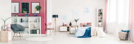 Grey armchair and pink wall in spacious bedroom interior with blue blanket on bed next to a wooden lampの写真素材