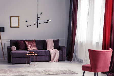 Red armchair and pink blanket on purple settee in bright living room interior with mockup of empty posterの写真素材