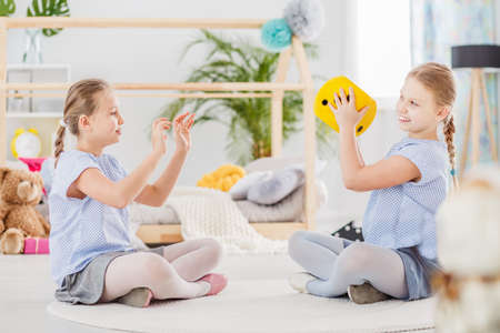 Smiling twin sisters playing with yellow plush dice in the classroomの写真素材
