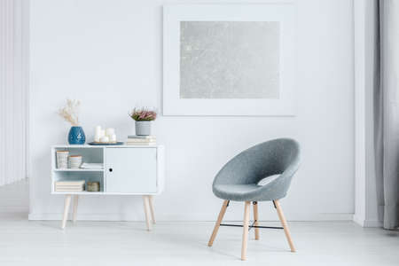 Grey chair next to white cupboard against the wall with silver painting in minimalist living room interiorの写真素材