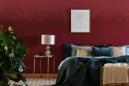 Lamp on a gold table next to green bed in luxurious bedroom interior with poster on red wallの写真素材