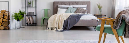 Green wooden chair with grey fur in spacious bedroom interior with ferns and knit blanket on bedの写真素材