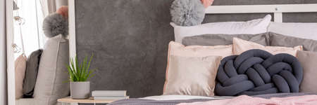 Knot pillow on bed next to a nightstand with books and aloe in cozy bedroom interior with mirrorの写真素材