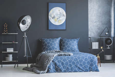 Industrial lamp next to bed with navy blue bedding against dark wall with moon poster in bedroom interiorの写真素材