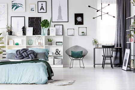 Freelancer's bedroom interior with blue bed and black, wooden chair at a desk near the window