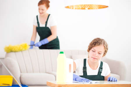 Young woman cleaning a wooden coffee table standing in a white living room