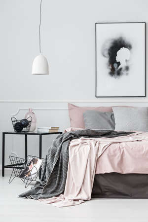 Black and white poster above bed with patterned blanket in bright bedroom interior with lampの写真素材