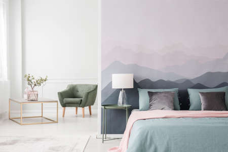 Large bed with blue sheets and a pink blanket by a landscape wallpaper in a cozy, modern bedroom interiorの写真素材