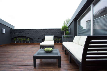 Wooden table and white garden furniture on terrace with black brick wall and view on the sky
