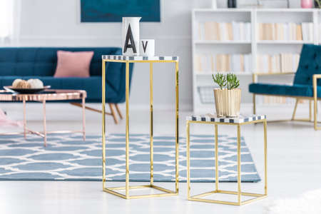 Close-up of gold tables with a plant and ceramic vessels with sofa and bookshelf in the background of living room interiorの写真素材
