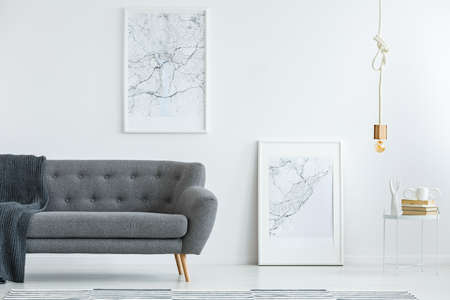 Elegant, gray sofa with wooden legs and large map posters on a white wall in a designer minimalist living room interior of an architectの写真素材