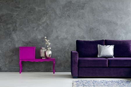 Violet cupboard with silver vases next to purple couch in dark living room interior with concrete...