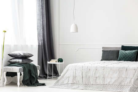 Silver pillows on bench and grey curtain in sophisticated bedroom interior with white lamp above bedの写真素材