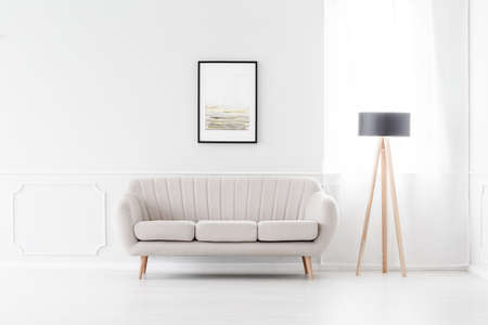Elegant, beige sofa in an empty white interior with art above the sofa and a wooden tripod lamp by a windowの写真素材