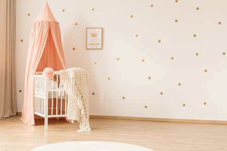 Classic white crib with a peach pink canopy in a girly nursery bedroom interior with gold polka dots...