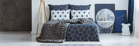 Knit wool blanket on patterned bed in bedroom interior with white table and moon poster