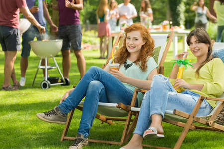 Close-up of two young attractive girls smiling at camera while sitting on deck chairs, enjoying hot summer day with drinks, garden barbecue party in the backgroundの写真素材