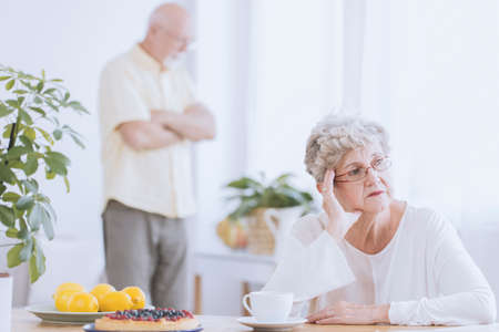 Elder woman sitting at a table, angry because of the argument with her husband standing behind herの写真素材