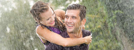 Smiling young man holding pretty girlfriend on his back during heavy summer rainの写真素材