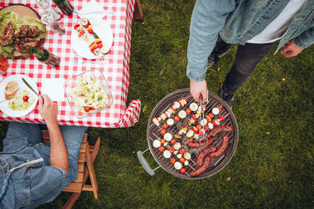 High angle of person grilling shashliks next to a man eating salad in the garden