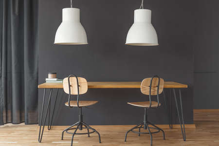 Two white lamps hanging above hairpin table in black dining room interior with curtain and chairsの写真素材