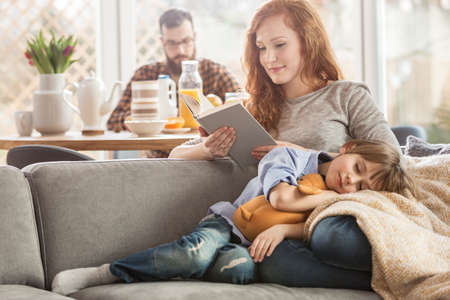 Sleeping son lying on mother's lap while she is reading a book and father eating in the backgroundの写真素材