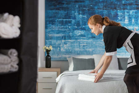 A young, working maid laying fresh, white towels on a bed in a hotel room