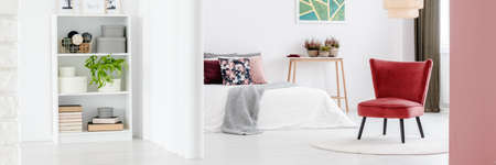 Photo of small rack with books, plants and decor and white bedroom interior with burgundy armchair...