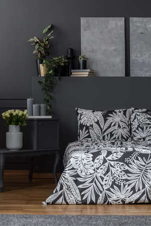 Grey paintings on dark wall above patterned bed in dark bedroom interior with flowersの写真素材