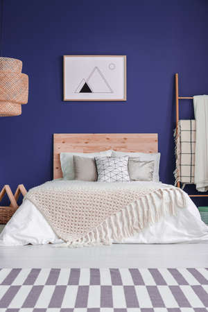 Poster on purple wall above bed with wooden bedhead and knit blanket in cozy bedroom interiorの写真素材