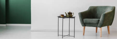 Grey armchair next to table with kettle against white wall in simple living room interiorの写真素材