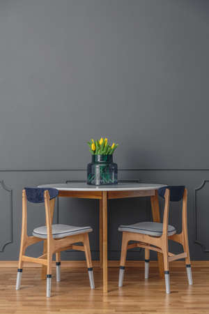 Paint-dipped chairs and a simple, wooden table with a bouquet of yellow tulips in glass vase by an empty, gray wall in a minimalist dining room interiorの写真素材