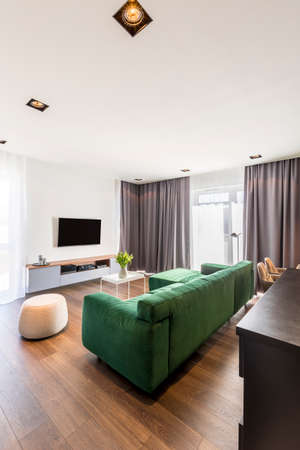 Spacious living room interior with green corner sofa, wooden floor, gray drapes and TV set