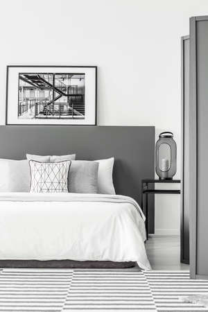 Black and white poster on bedhead of bed in simple bedroom interior with lantern on a bedside tableの写真素材