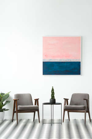 Plant on a table between grey armchairs in modern interior with pink and blue painting on white wall
