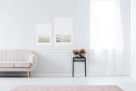 Heathers on black table near sofa against white wall with posters in living room interior with...