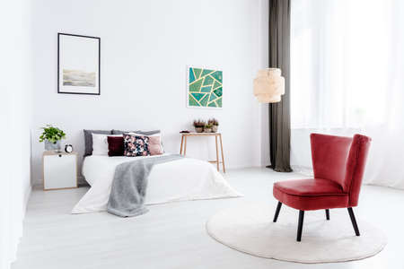 Red armchair on round rug in white bedroom interior with green painting and poster on the wallの写真素材