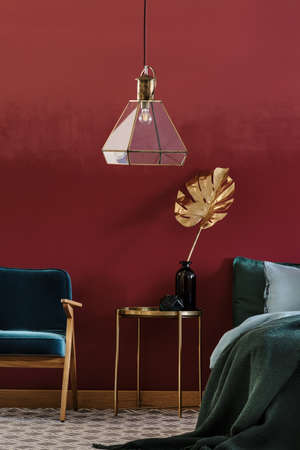 Golden, decorative leaf in glass vase standing on a metal bedside table between blue armchair and bed in bedroom interior の写真素材