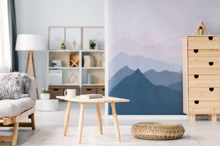 Blurred photo of a scandi living room interior with mountain wallpaper and beige sofa. Focus on a wooden table and pouf in the middle.の写真素材