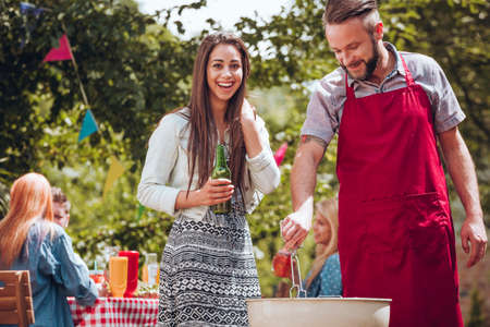 Cheerful young couple standing by a grill, the woman holding a green bottle of beer, the man holding cooking tongs at a summer garden partyの写真素材