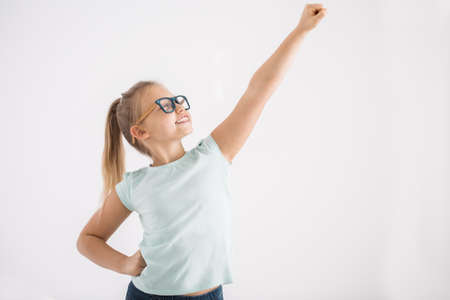 Portrait of a young girl with her arm and fist up and the other arm on a hip in a superhero pose on white wall backgroundの写真素材