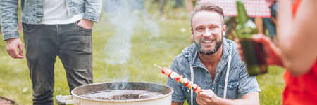 Smiling man grilling shashlik during barbeque party with friendsの写真素材