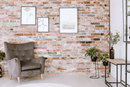 Living room interior with grey armchair, plants and posters on a red brick wall 
