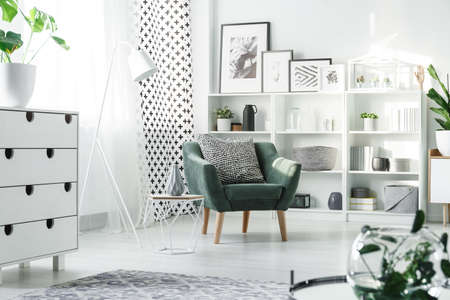 White furniture and green armchair with patterned pillow placed in bright studio interior with...