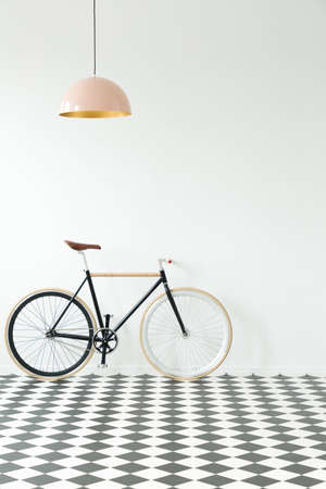 Black bike against white wall in simple interior with checkerboard floor and pink lampの写真素材