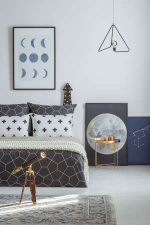 Telescope on patterned carpet and copper table next to bed in bedroom interior with moon poster