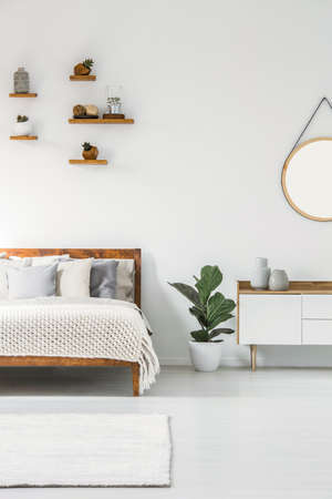 Plant between white cupboard and bed in scandi bedroom interior with round mirrorの写真素材
