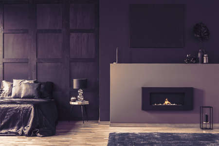 Fireplace in elegant dark bedroom interior with lamp on wooden table next to bedの写真素材