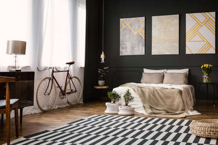 Modern bedroom interior with black wall, bike by the window, lamp, chair, pouf, plants, paintings and carpetの写真素材