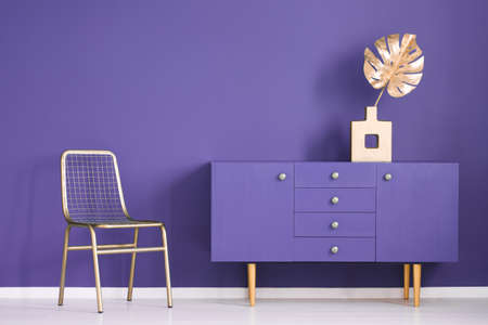 Golden monstera leaf in a vase standing on an ultra violet cupboard next to a metal chair in creative living room interior. Real photoの写真素材